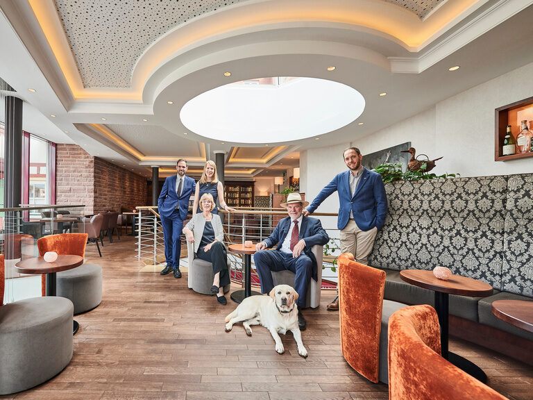 Familie Berlin mit Hund Prinz, stehend und sitzend auf den modernen, orangenen und grauen Sesseln, in der Lounge.