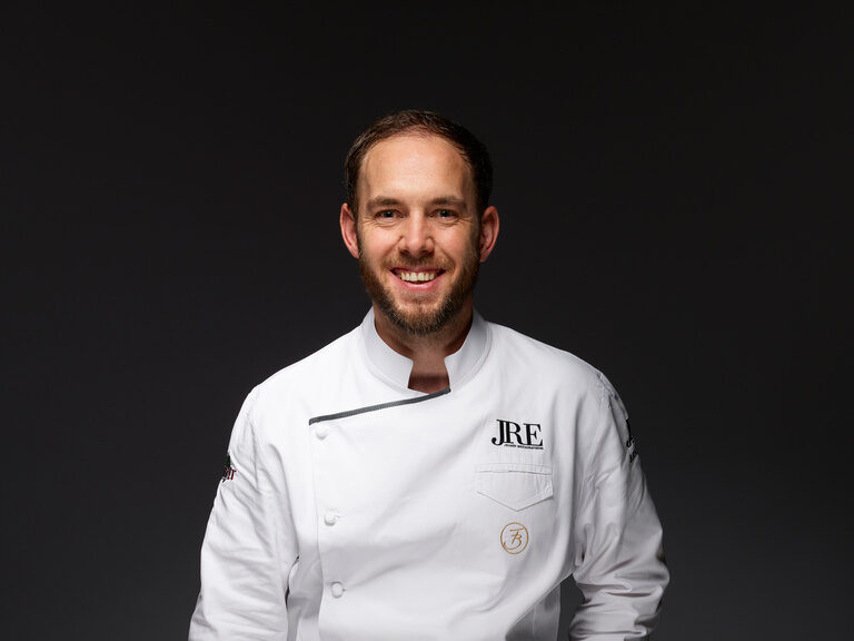 Franz Berlin, in weißer Kochjacke, lächelnd vor einem dunklen Hintergrund.