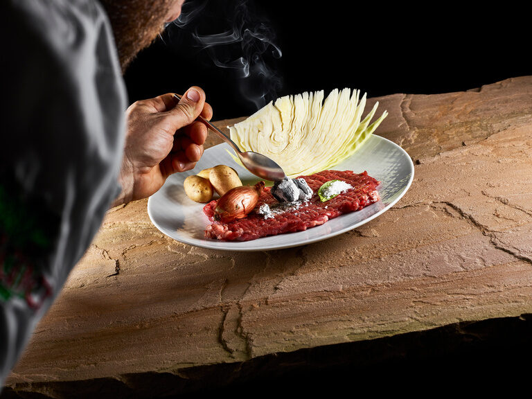 Auf einem Teller liegen ein Stück geklopftes Rindfleisch mit Kartoffeln, Weißkraut sowie Zwiebeln.