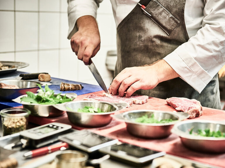 Ein Koch schneidet Fleisch auf einem Brett vor ihm stehen diverse Küchenutensilien sowie verschiedene Kräuter.